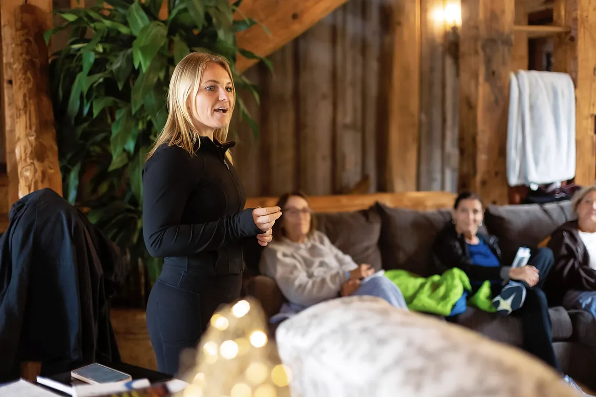 Lacey Cook stands and speaks to participants seated on sofas in a wood paneled living room.